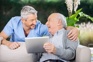 An older patient holding a tablet and sitting on the couch while a doctor stands over him with his arm around him